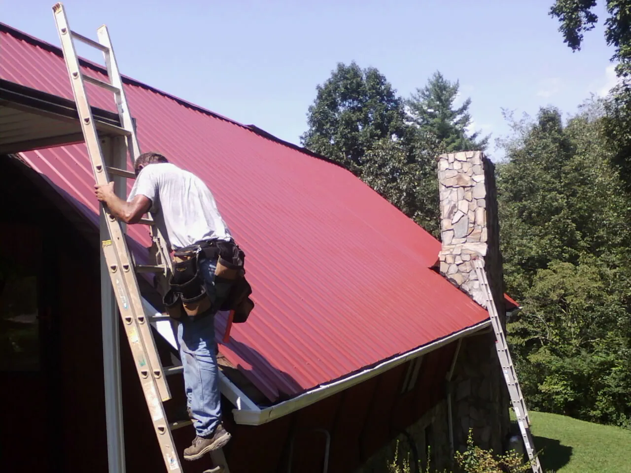 Roofer on a ladder beside a red metal roof and stone chimney — Soffit & Fascia Repair by Roofing Brothers in Kenansville